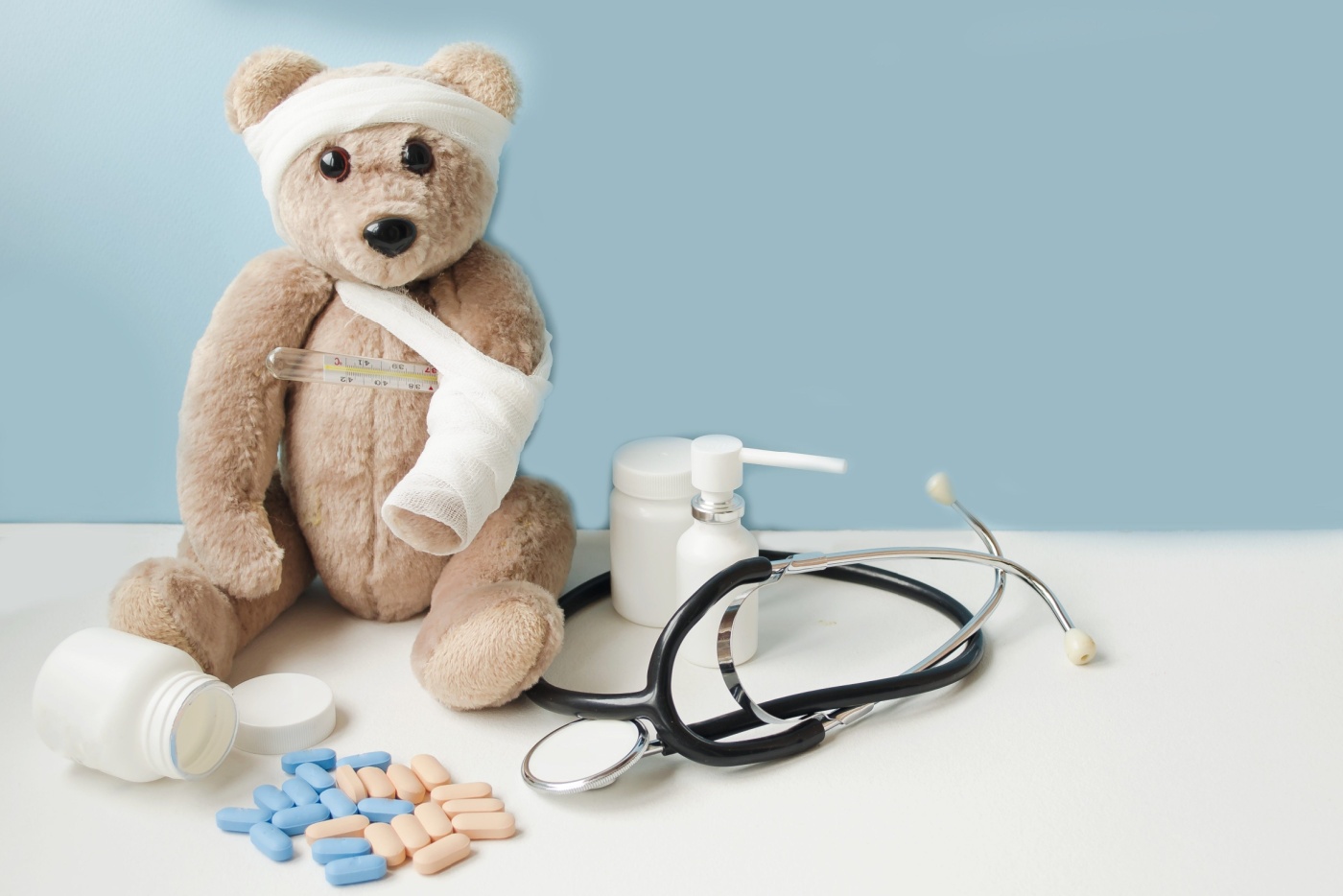 Foto: Shutterstock, En bamse med brukket pote og vondt hode sitter på blå bakgrunn, ved siden av piller, vitaminer og et stetoskop