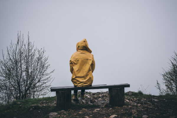 Fotografi av person sittende med ryggen til på en benk, i regnvær