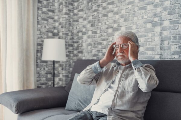 Fotografi av eldre mann i sofa, som tar seg til hodet. Frustrert, mentalt sliten.