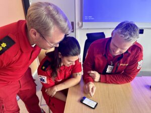 Bilde viser tre ambulansearbeidere, der de ser på appen det står om i artikkelen på en mobiltelefon.