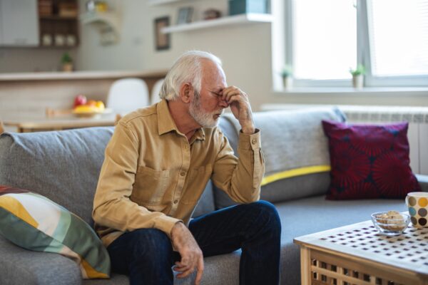 Fotografi av senior som tar seg til hodet, pga svimmelhet, sittende på en sofa